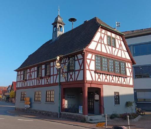 Fachwerkhaus mit rotem Gebälk, weißen Wänden und einem kleinen Turm auf dem Dach, aufgenommen von der Ostseite bei blauem Himmel.