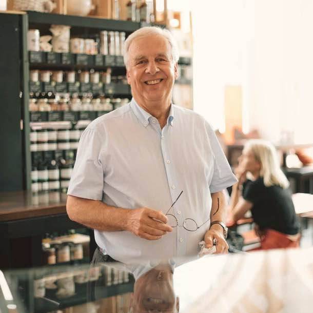 Älterer Mann mit grauen Haaren und hellem Hemd steht lächelnd in einem Laden vor einem Regal mit Gläsern und Gewürzen, hält eine Brille in der Hand. Im Hintergrund sitzt eine Frau an einem Tisch.
