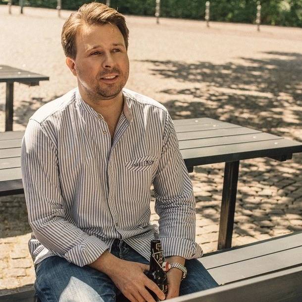 Ein Mann sitzt auf einer Parkbank im Freien, hält eine Flasche in der Hand und trägt ein gestreiftes Hemd.
