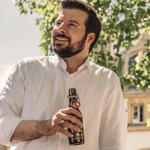 Ein Mann mit Bart und weißem Hemd steht draußen, hält eine Flasche Bier in der Hand und lächelt. Im Hintergrund sind grüne Bäume und ein Gebäude zu sehen.
