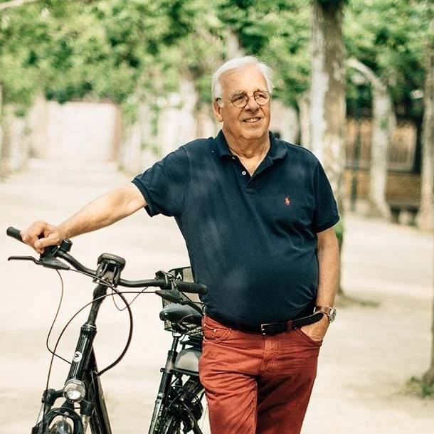 Älterer Mann mit grauen Haaren und Brille steht entspannt im Freien neben einem Fahrrad, trägt ein dunkelblaues Polohemd und rote Hose. Im Hintergrund sind Bäume und ein Weg zu sehen.
