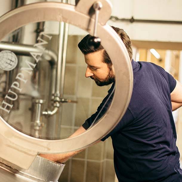 Ein Mann steht in einer Brauerei und arbeitet an einem großen Edelstahl-Braukessel mit der Aufschrift 