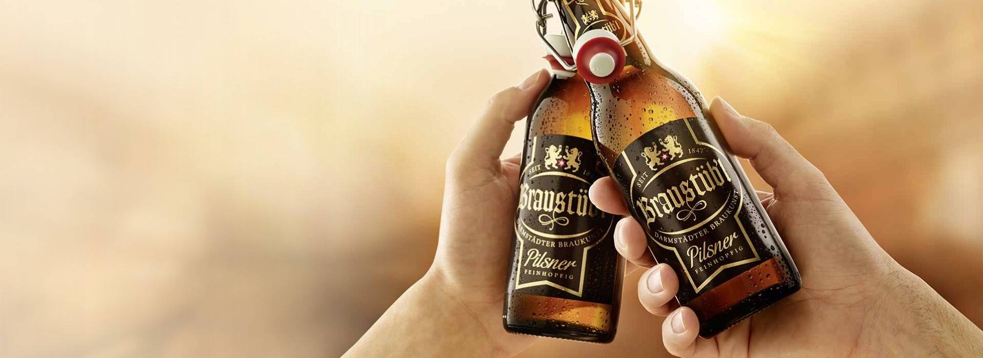 Zwei Hände stoßen mit Braustübl Pilsner-Bierflaschen vor unscharfem Hintergrund an.