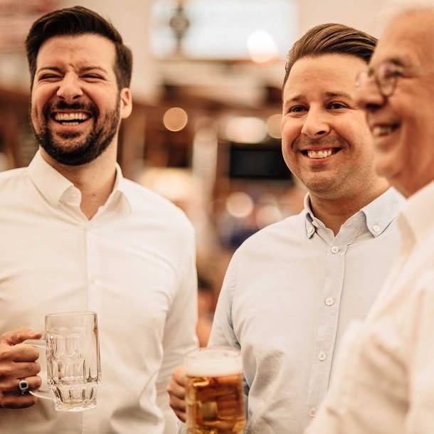 Drei lachende Männer in weißen Hemden halten Biergläser in der Hand und stehen in geselliger Runde.