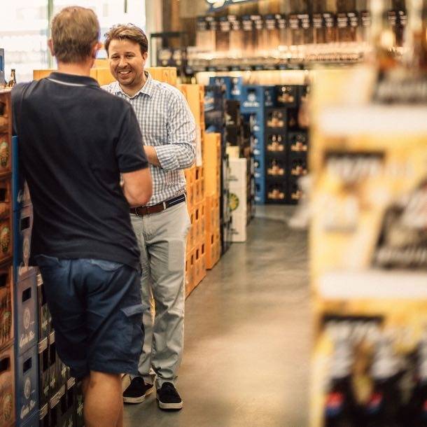 Zwei Männer unterhalten sich lachend in einem Getränkemarkt zwischen Getränkekisten.