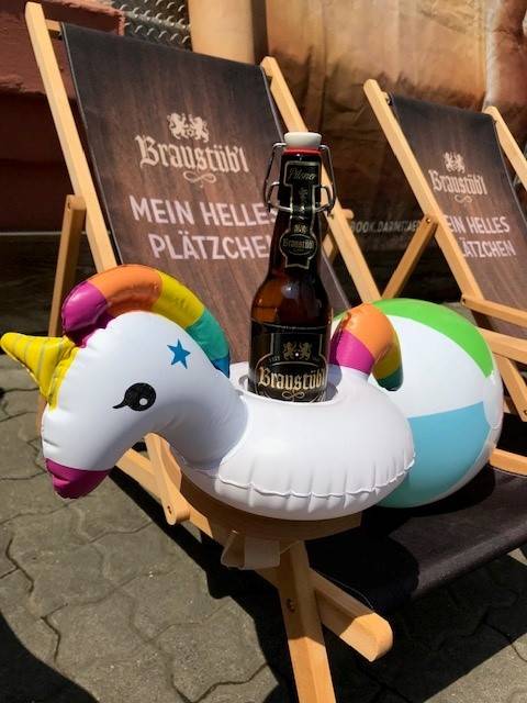 Eine Flasche Braustübl-Bier steht in einem aufblasbaren Einhorn-Getränkehalter auf einem Liegestuhl, daneben ein bunter Wasserball; im Hintergrund zwei weitere Liegestühle mit der Aufschrift „Braustübl – Mein helles Plätzchen“.