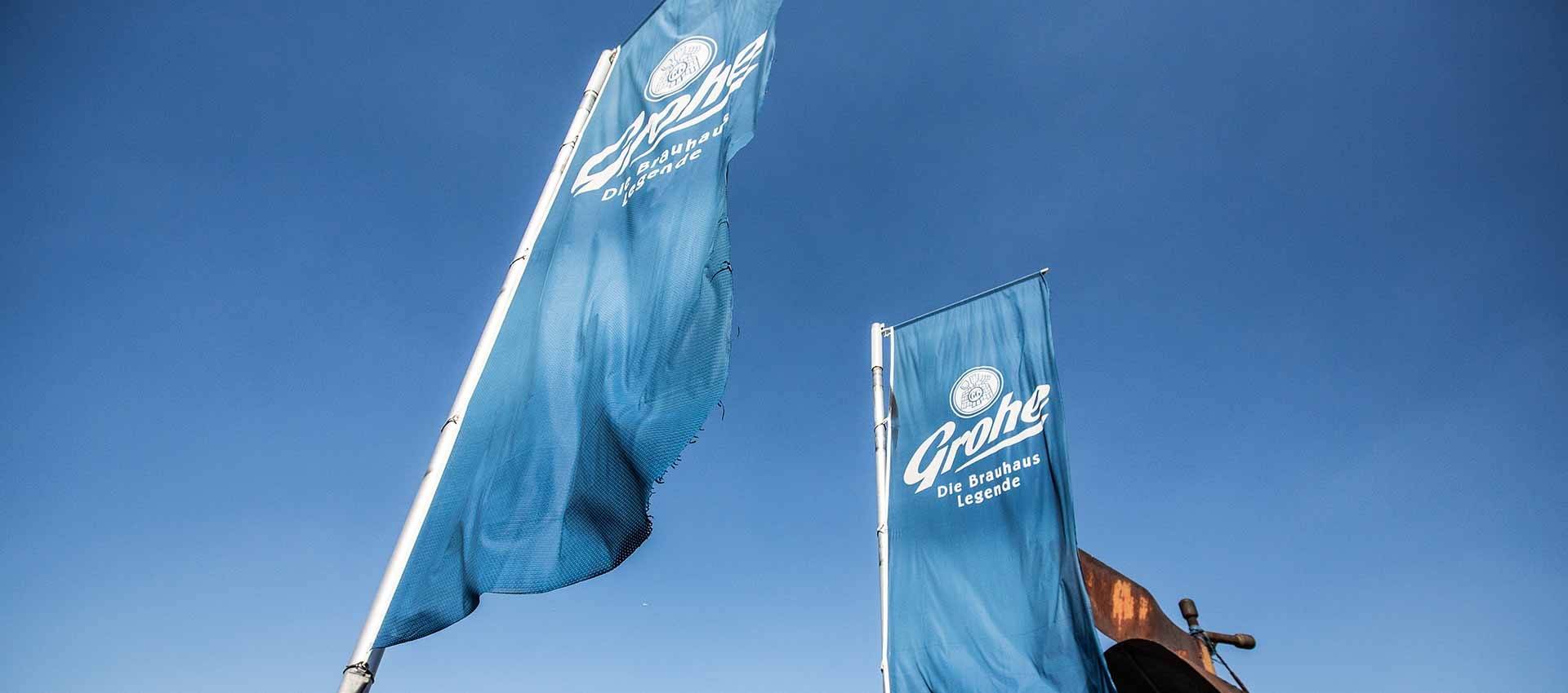 Zwei blaue Fahnen mit weißem Schriftzug „Grohe Die Brauhaus Legende“ wehen im Wind vor blauem Himmel.