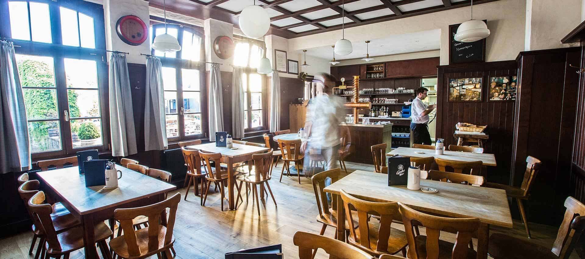 Helles, traditionelles Wirtshaus mit Holztischen und -stühlen, großen Fenstern, Zapfanlage an der Theke und zwei Bedienungen.