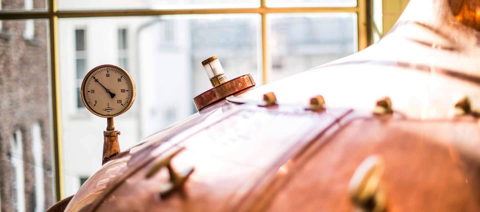 Nahaufnahme eines alten, kupfernen Braukessels mit einem analogen Temperatur- oder Druckmesser vor einem Fenster.