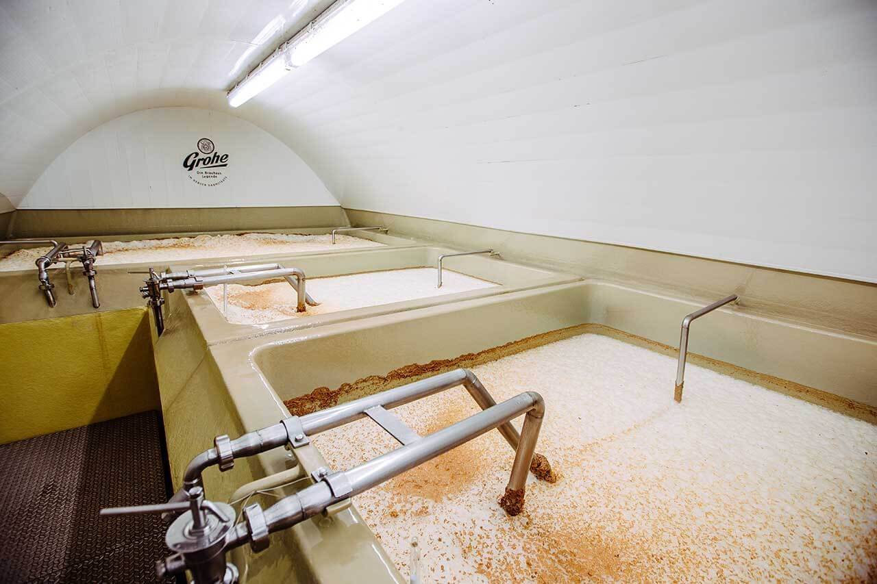 Großer Raum mit mehreren rechteckigen Gärbottichen, gefüllt mit schäumender Flüssigkeit und Rohren, an der Wand das Logo „Grohe“.