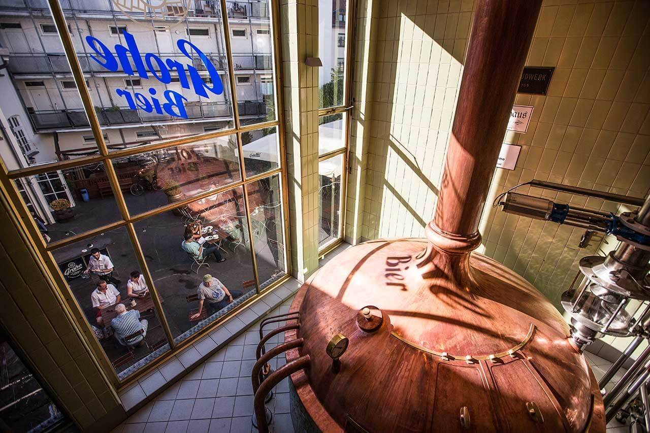 Großer Kupferkessel in einer Brauerei, Blick durch ein Fenster auf einen Außenbereich, wo mehrere Menschen an Tischen sitzen; auf dem Fenster steht „Grohe Bier“.