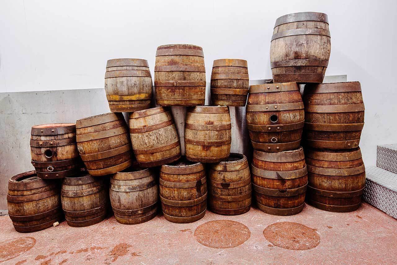 Mehrere alte, leere Holzfässer sind in einer Ecke aufeinander gestapelt; der Boden ist rot, die Wand weiß.