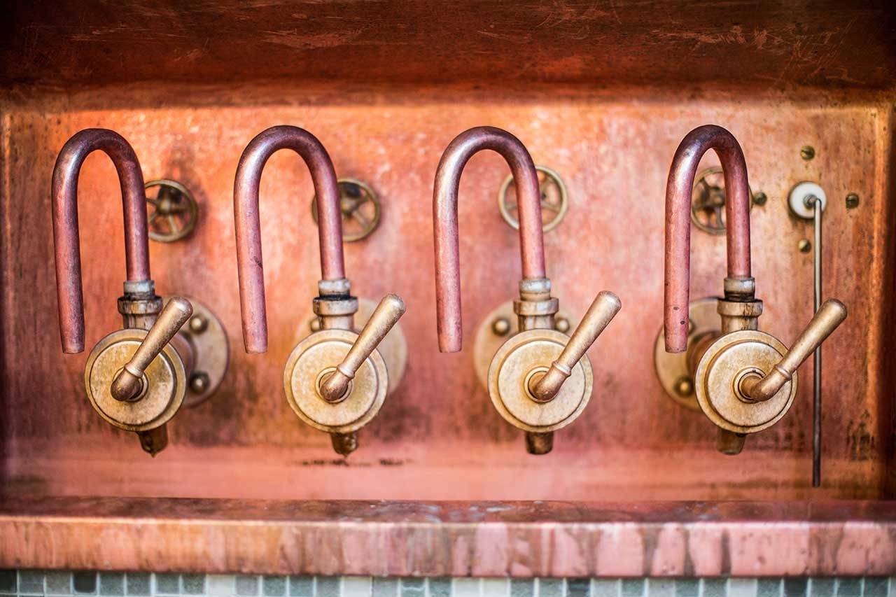 Vier alte, messingfarbene Wasserhähne mit gebogenen Kupferrohren an einer rötlichen Wand.