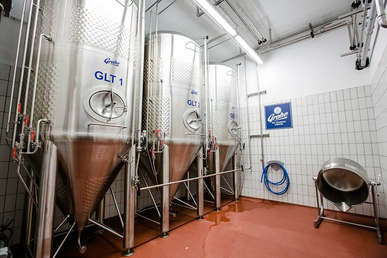 Mehrere große Edelstahltanks in einem Brauerei-Raum mit gefliestem Boden und Wänden, daneben ein blauer Wasserschlauch und ein Metallschild mit der Aufschrift 