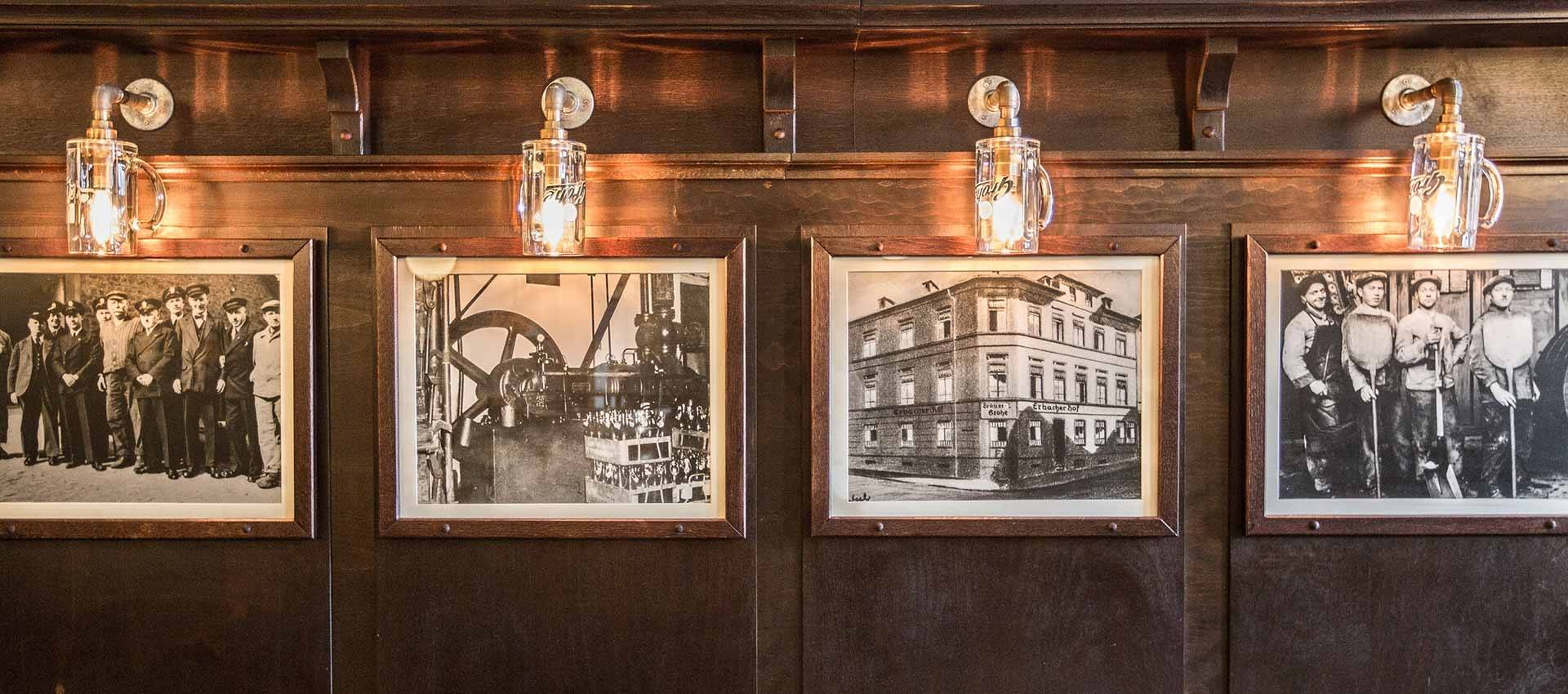 Vier gerahmte Schwarzweiß-Fotografien an einer dunklen Holzwand, beleuchtet von Lampen in Form von Bierkrügen. Die Bilder zeigen historische Gruppen von Menschen, eine alte Maschine und ein Gebäude.