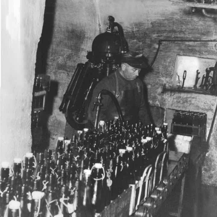 Schwarzweißfoto eines Mannes in Arbeitskleidung, der in einem Kellerraum zwischen vielen Glasflaschen steht und anscheinend eine Abfüllanlage bedient.