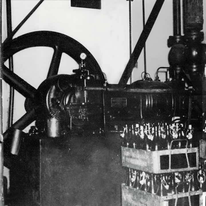 Schwarzweißfoto einer alten industriellen Maschine mit großem Schwungrad im Hintergrund; im Vordergrund stapeln sich Holzkisten mit Glasflaschen.
