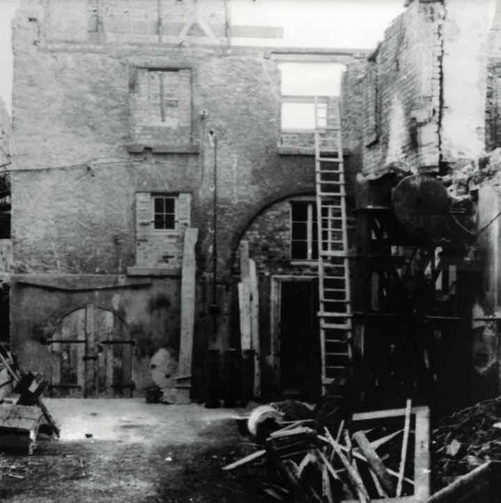 Schwarz-weiß-Foto eines teilweise zerstörten Gebäudes mit offenen Wänden, einer Leiter und herumliegendem Schutt.
