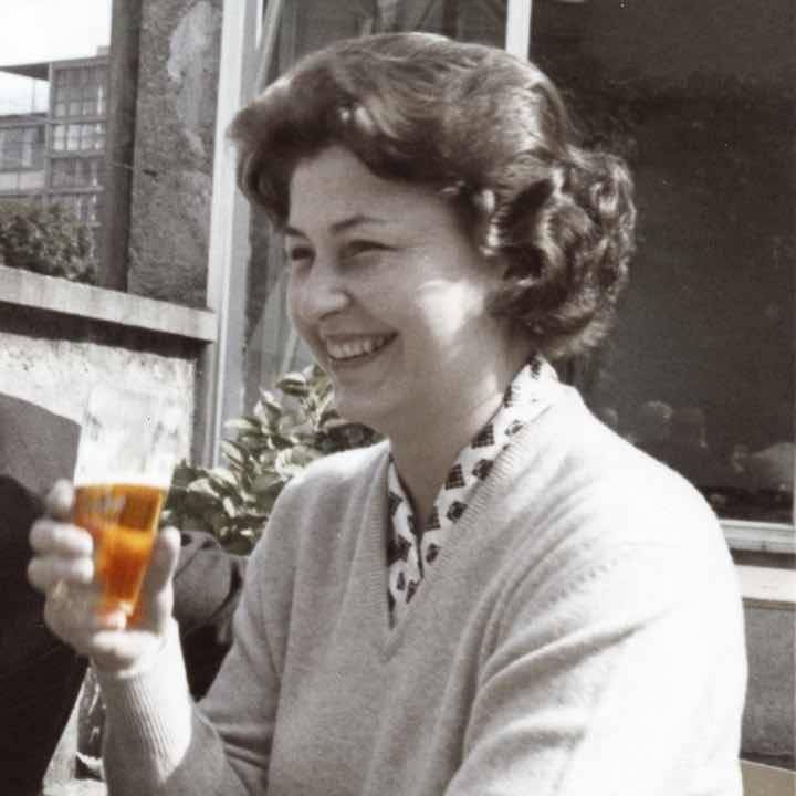 Schwarz-weiß-Foto einer lächelnden Frau mit welligen, kurzen Haaren, die im Freien ein Glas mit einem Getränk in der Hand hält.