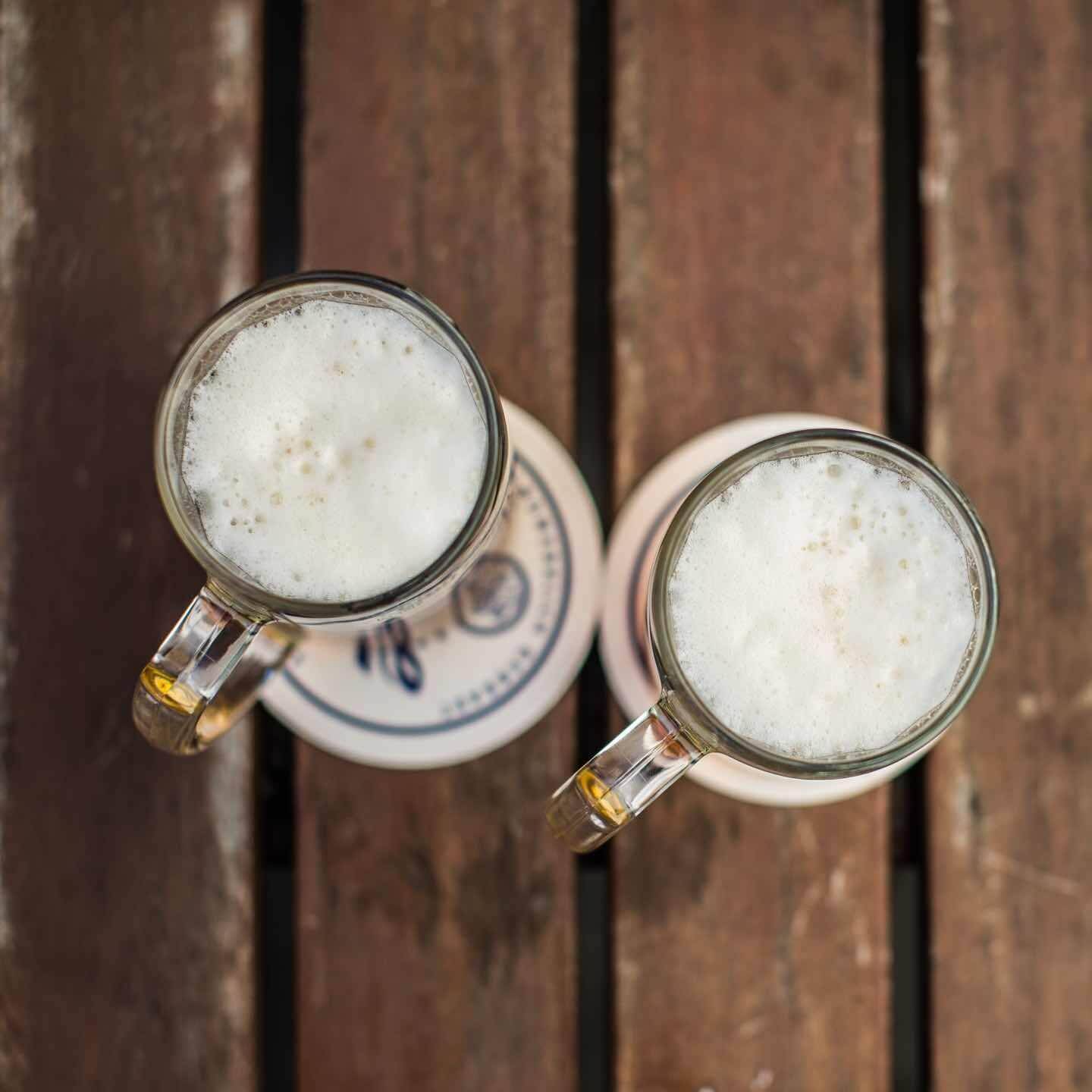 Zwei Bierkrüge mit Schaum stehen nebeneinander auf Bierdeckeln auf einem Holztisch, von oben fotografiert.