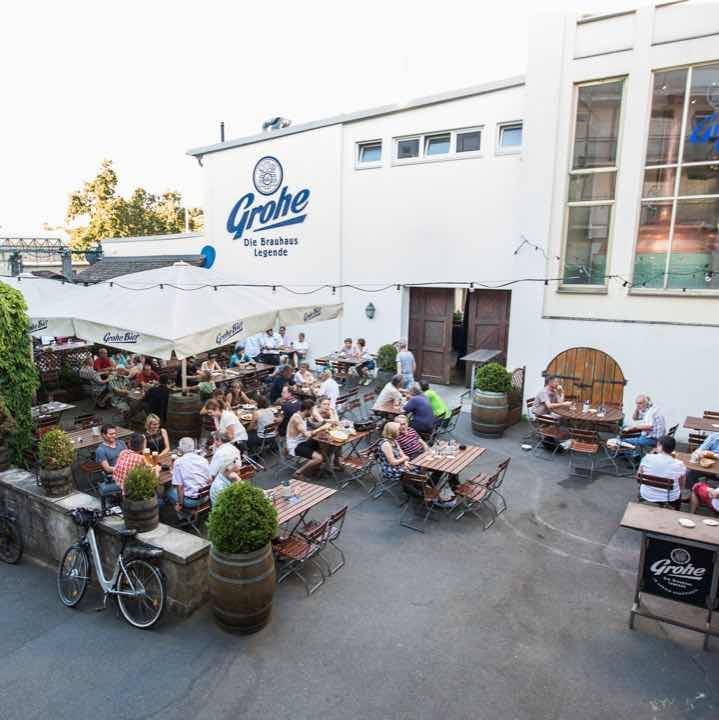 Außenbereich einer Brauerei mit mehreren Holztischen und Bänken, an denen Menschen sitzen, essen und trinken; im Hintergrund ein Gebäude mit dem Schriftzug 