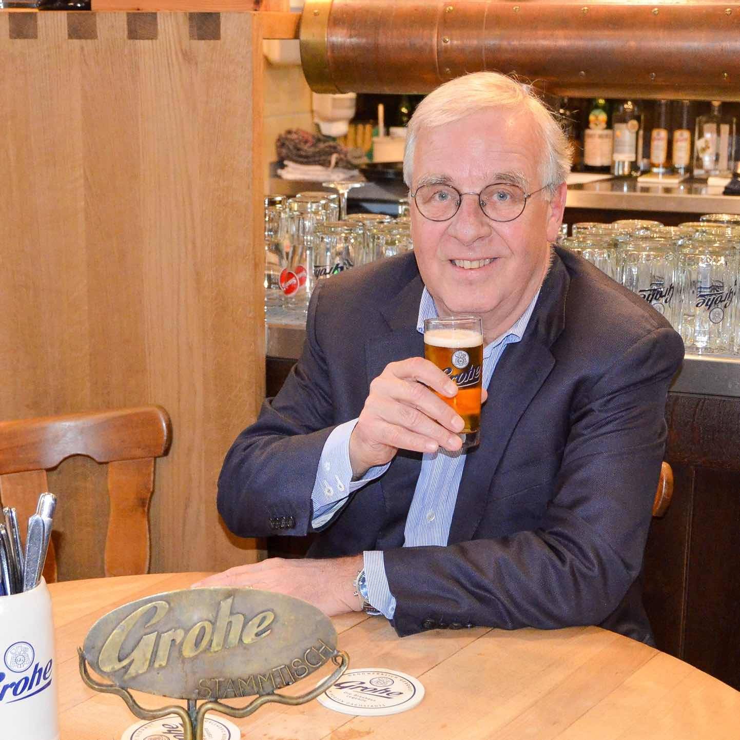 Ein älterer Mann mit Brille und grauem Haar sitzt an einem Holztisch in einer Kneipe, hält ein Glas Bier in der Hand und lächelt in die Kamera. Auf dem Tisch steht ein Metallschild mit der Aufschrift „Grohe Stammtisch“. Im Hintergrund sind viele Gläser und eine Bar zu sehen.