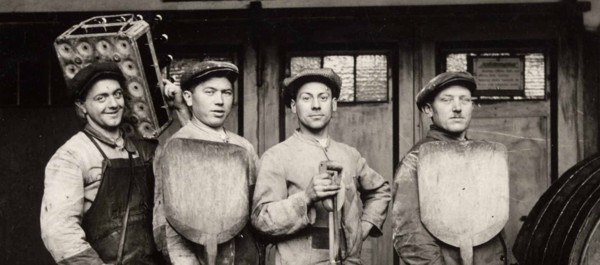 Schwarzweißfoto von vier Bäckern mit Schiebern vor einer Bäckerei, alle tragen Arbeitskleidung und Mützen.