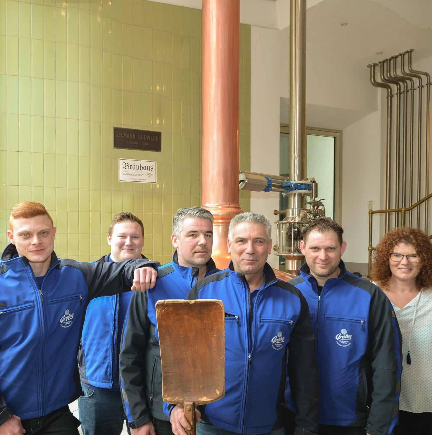 Sechs Personen in blauer Arbeitskleidung stehen in einer Brauerei vor einer kupfernen Brauanlage; eine Person hält eine hölzerne Schaufel.