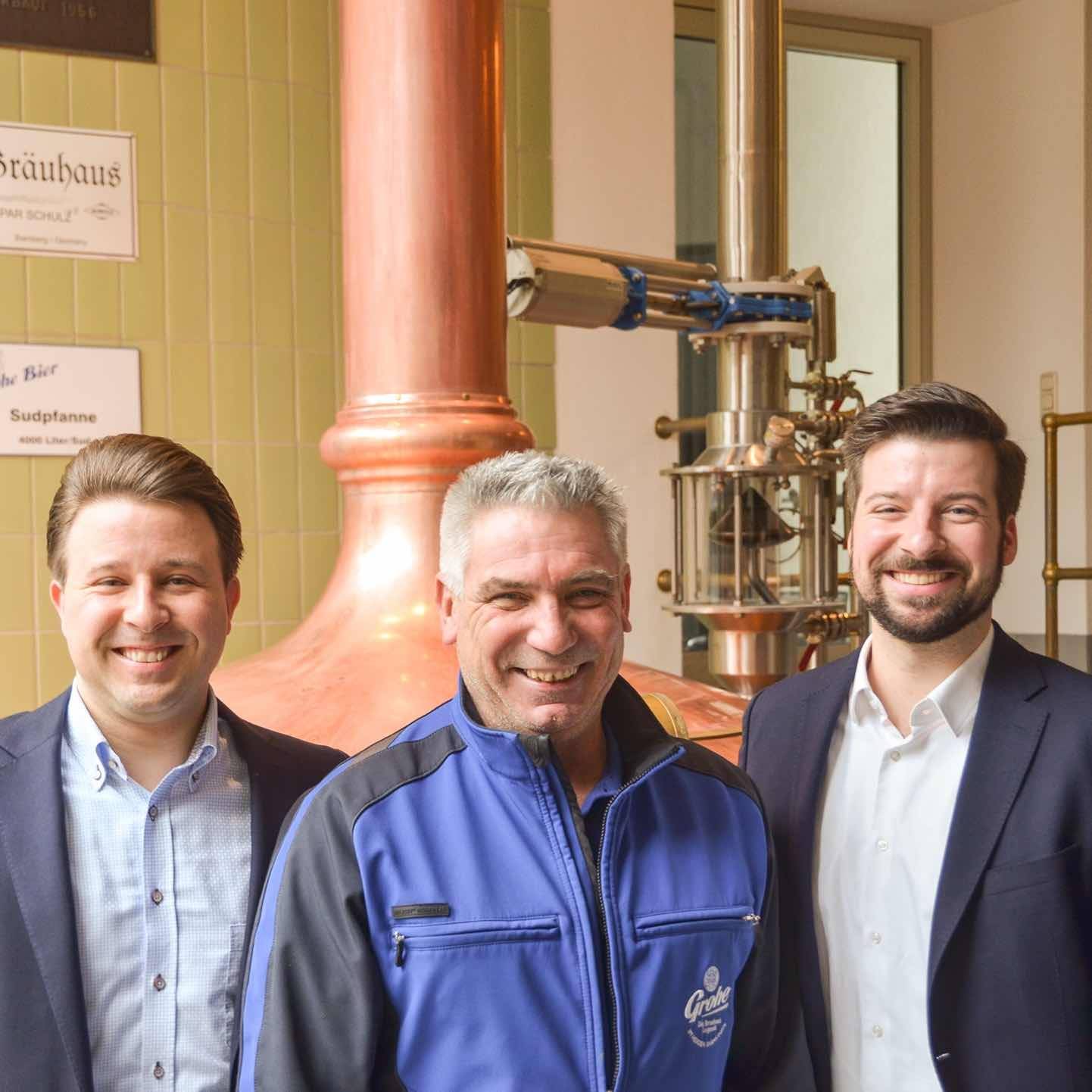 Drei lächelnde Männer stehen vor einer großen Kupferbrauanlage in einer Brauerei.