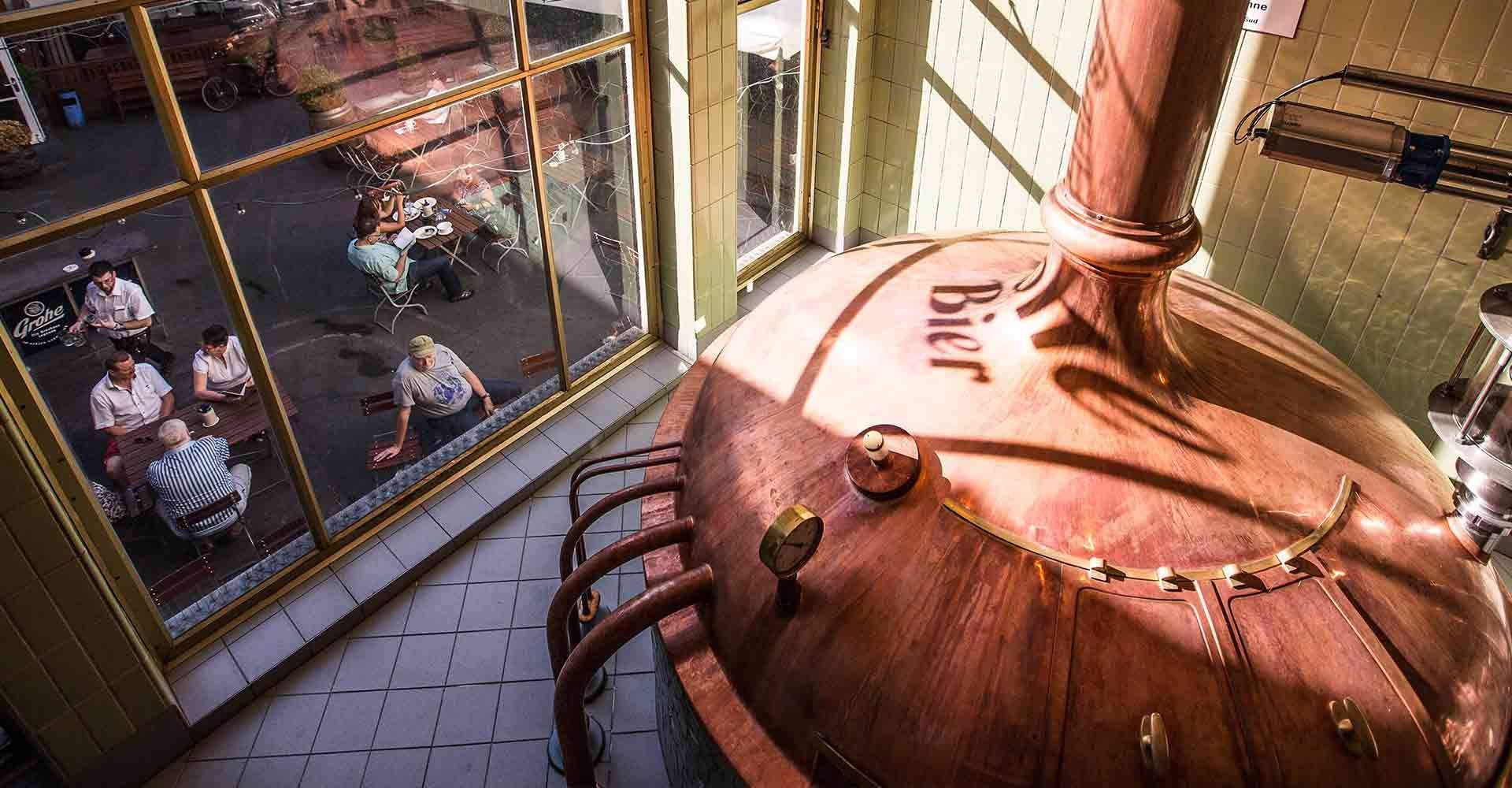 Großer Braukessel in einem Brauhaus, daneben Fenster mit Blick auf draußen sitzende Menschen an Tischen.
