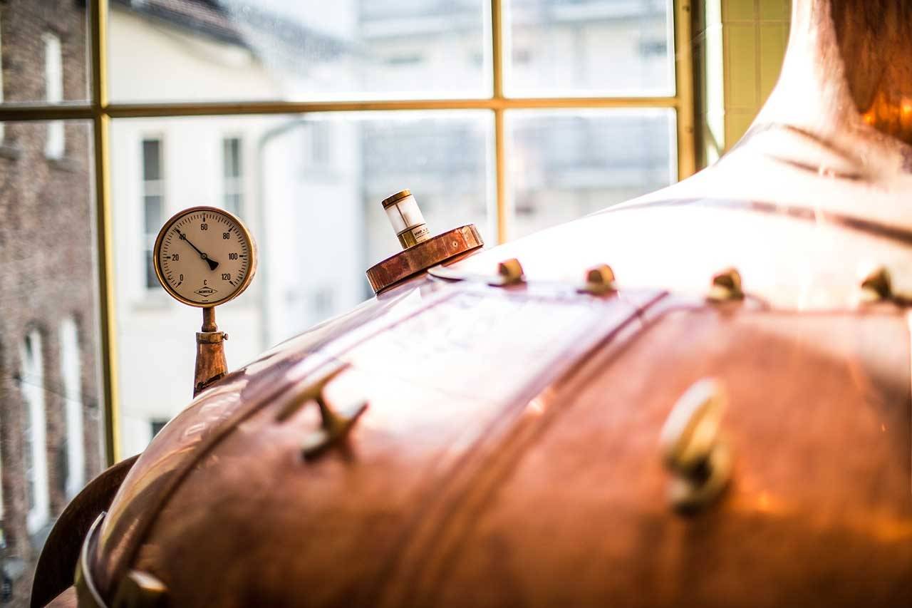 Großer Kupferkessel mit Manometer und Armaturen, im Hintergrund ein Fenster mit unscharfer Außenansicht.