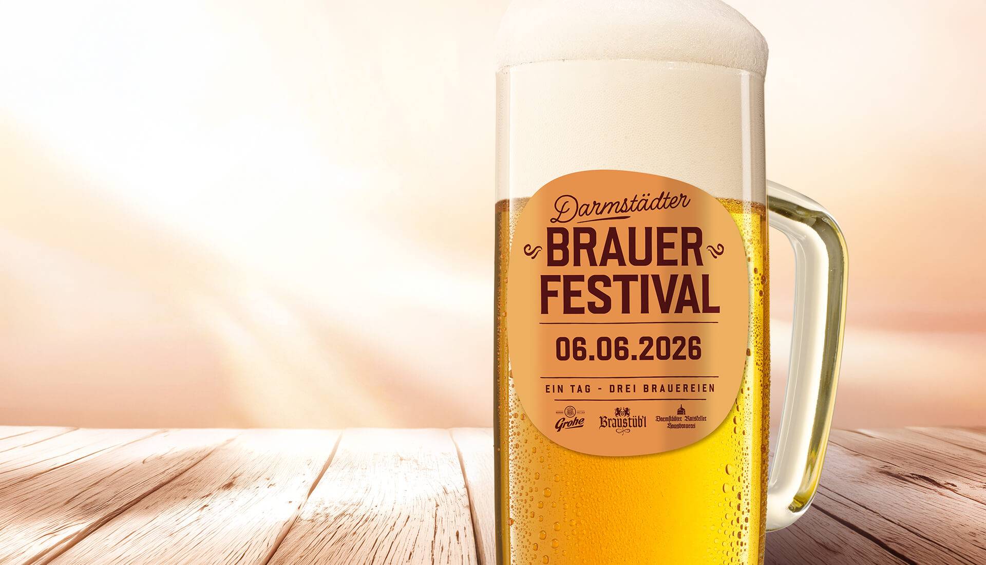 Ein großes Bierglas mit Schaumkrone steht auf einem Holztisch, darauf ein Etikett mit der Aufschrift „Darmstädter Brauer Festival, 06.06.2026, Ein Tag – Drei Brauereien“. Im Hintergrund ein heller, unscharfer Verlauf.