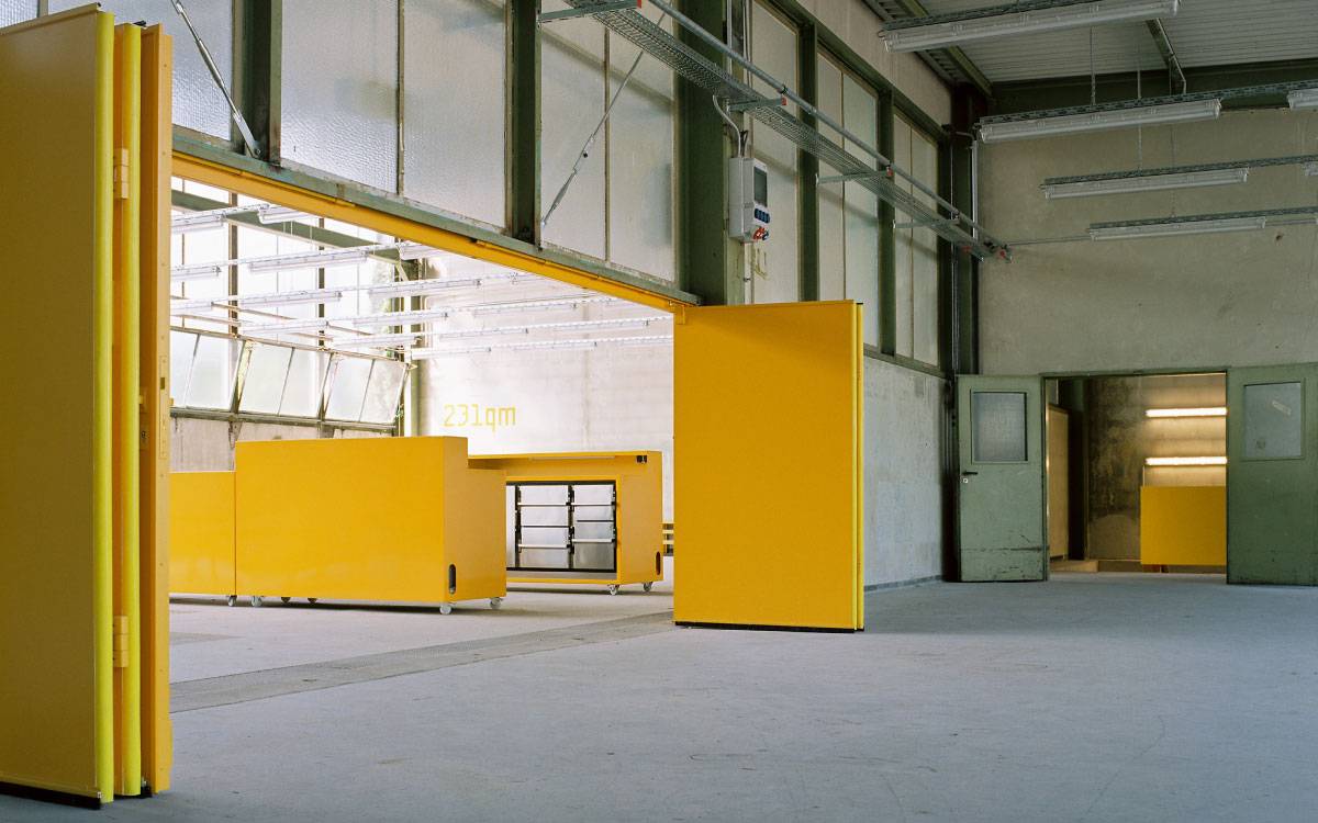 Großer, leerer Industrie- oder Lagerraum mit offenen, gelben Schiebetüren und gelben Möbeln auf Rollen, viel Tageslicht durch Fenster.