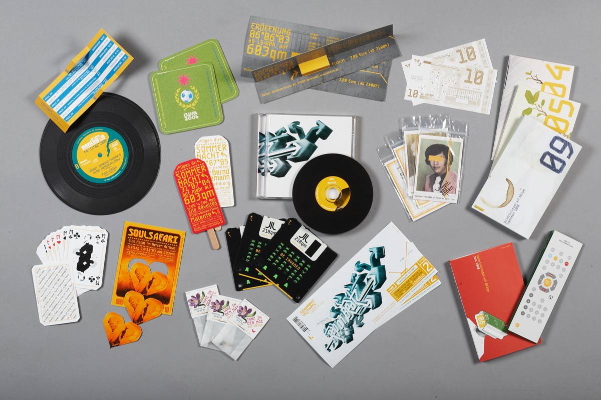Verschiedene Werbematerialien und Druckprodukte wie Schallplatten, Flyer, Eintrittskarten, Spielkarten, Bierdeckel, CD, Postkarten und ein Fernsehfernbedienungsmodell sind auf einem grauen Untergrund ausgebreitet.