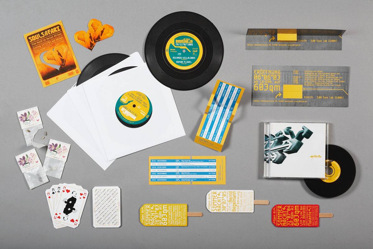 Verschiedene Designobjekte wie Schallplatten, Flyer, Spielkarten, Eisstiele mit Text, Teebeutel, eine CD und Tickets sind flach auf grauem Hintergrund arrangiert.