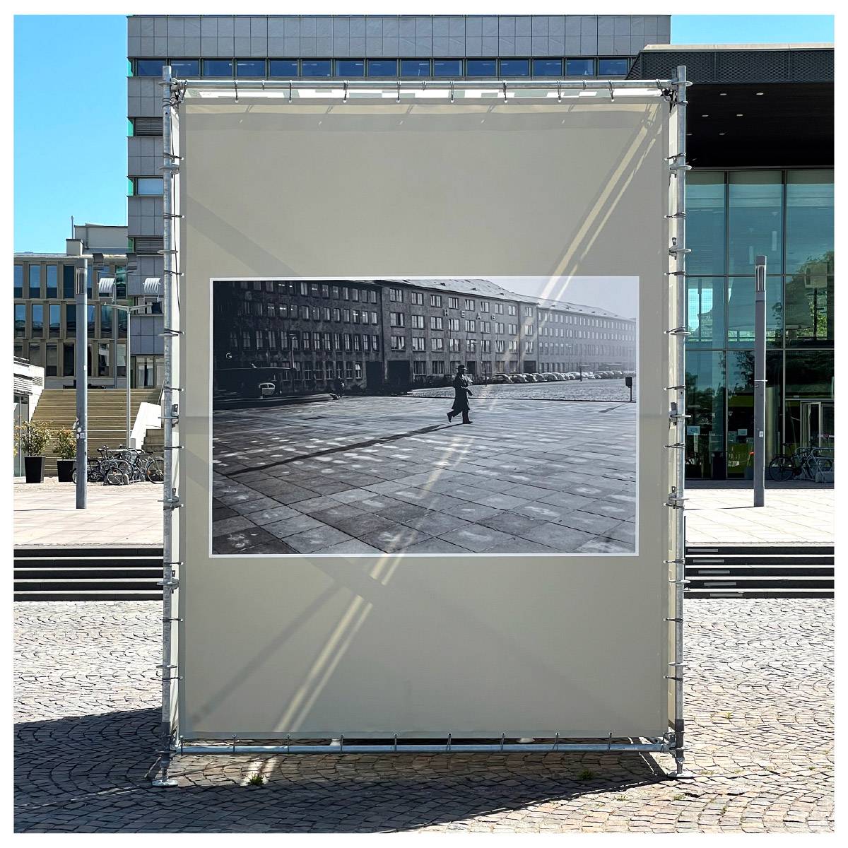 Große Outdoor-Ausstellungstafel mit einem schwarz-weißen Foto einer einzelnen Person, die über einen gepflasterten Platz vor einem langen Gebäude geht; moderne Gebäude im Hintergrund.
