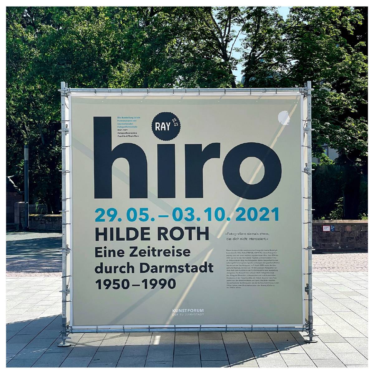 Großes Plakat im Freien mit der Aufschrift: „hiro – Hilde Roth. Eine Zeitreise durch Darmstadt 1950–1990. 29.05. – 03.10.2021“ vor grünem Baumbestand.