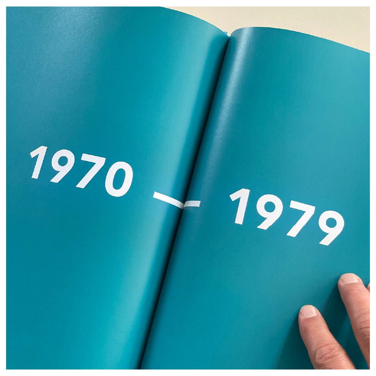 Aufgeschlagenes Buch mit türkisfarbenen Seiten, darauf in Weiß die Zahlen „1970 – 1979“, unten rechts eine Hand.