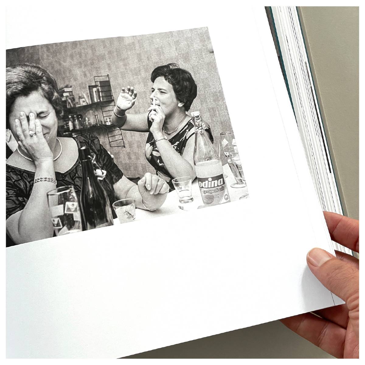 Schwarzweißfoto von zwei lachenden Frauen an einem Tisch mit Getränkeflaschen und Gläsern, eine Frau hält sich die Hand vors Gesicht, die andere gestikuliert lachend.