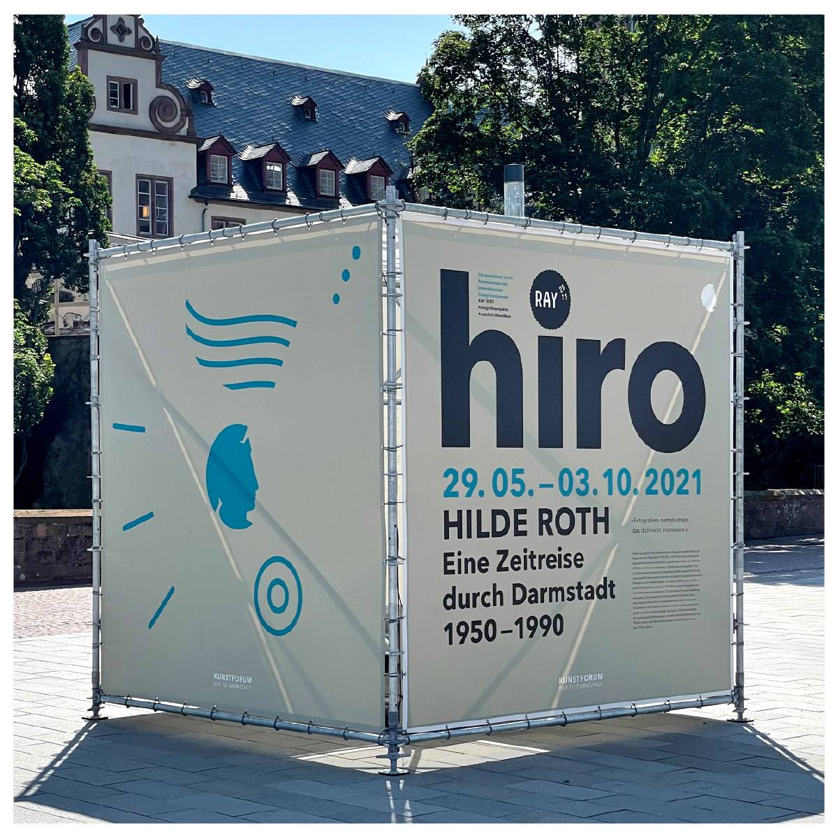 Großer, würfelförmiger Aufsteller im Freien mit der Aufschrift: "hiro, 29.05.–03.10.2021, Hilde Roth, Eine Zeitreise durch Darmstadt 1950–1990" sowie grafischen Elementen und einem stilisierten Profilkopf. Im Hintergrund Bäume und ein historisches Gebäude.