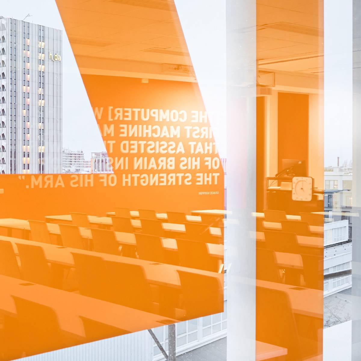 Blick durch eine Glaswand mit orangefarbenen Streifen in einen leeren, modernen Hörsaal; im Hintergrund ist ein Hochhaus mit der Aufschrift "h da" zu sehen.