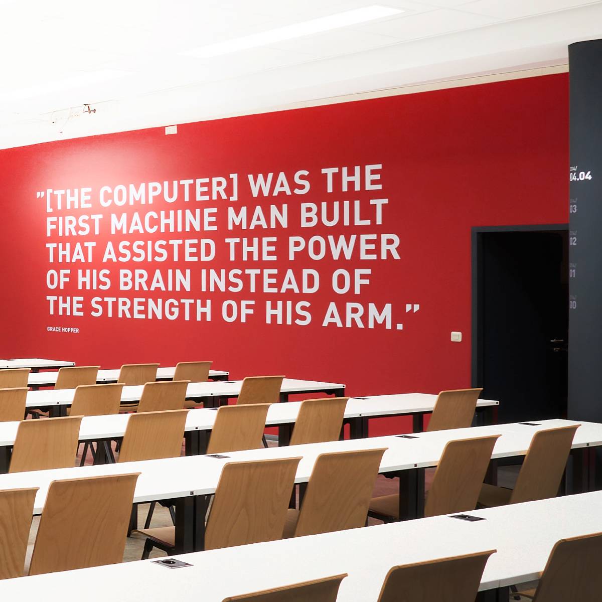 Klassenzimmer mit weißen Tischen und Holzstühlen; an einer roten Wand steht ein großes Zitat von Grace Hopper über Computer.