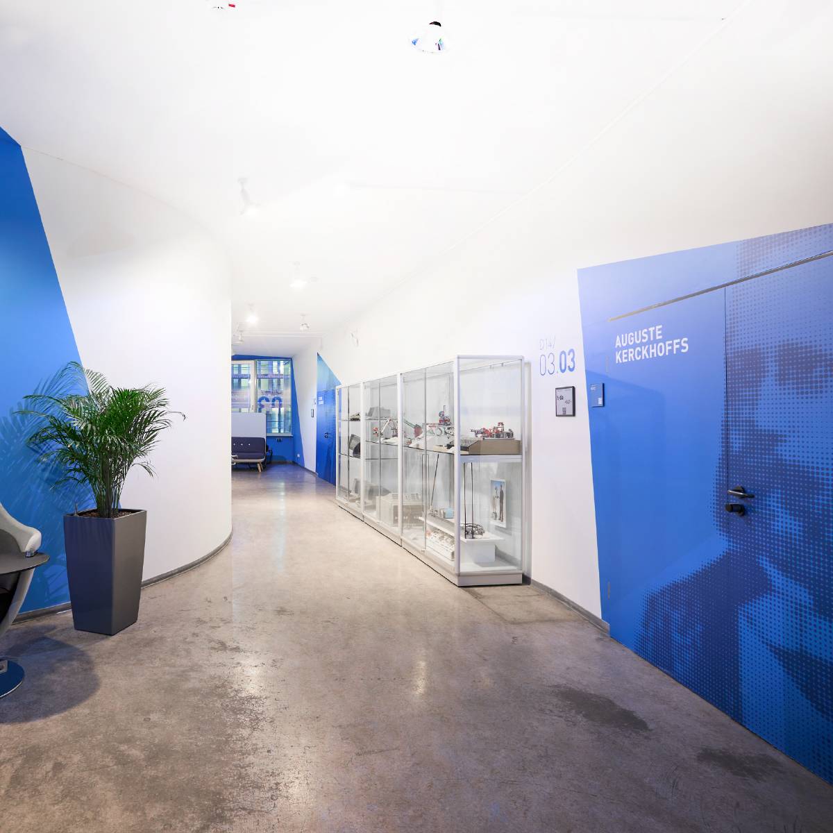 Heller, moderner Flur mit blauen Akzenten, einer Glastür mit dem Namen „Auguste Kerckhoffs“, Vitrine mit Ausstellungsstücken und einer großen Topfpflanze.