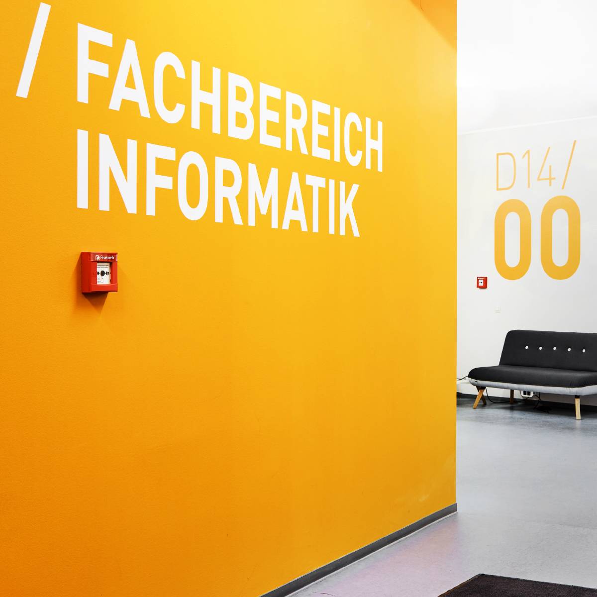 Gelbe Wand mit weißer Aufschrift „Fachbereich Informatik“, daneben ein roter Feuermelder, im Hintergrund eine schwarze Sitzbank und die Raumbezeichnung „D14/00“.