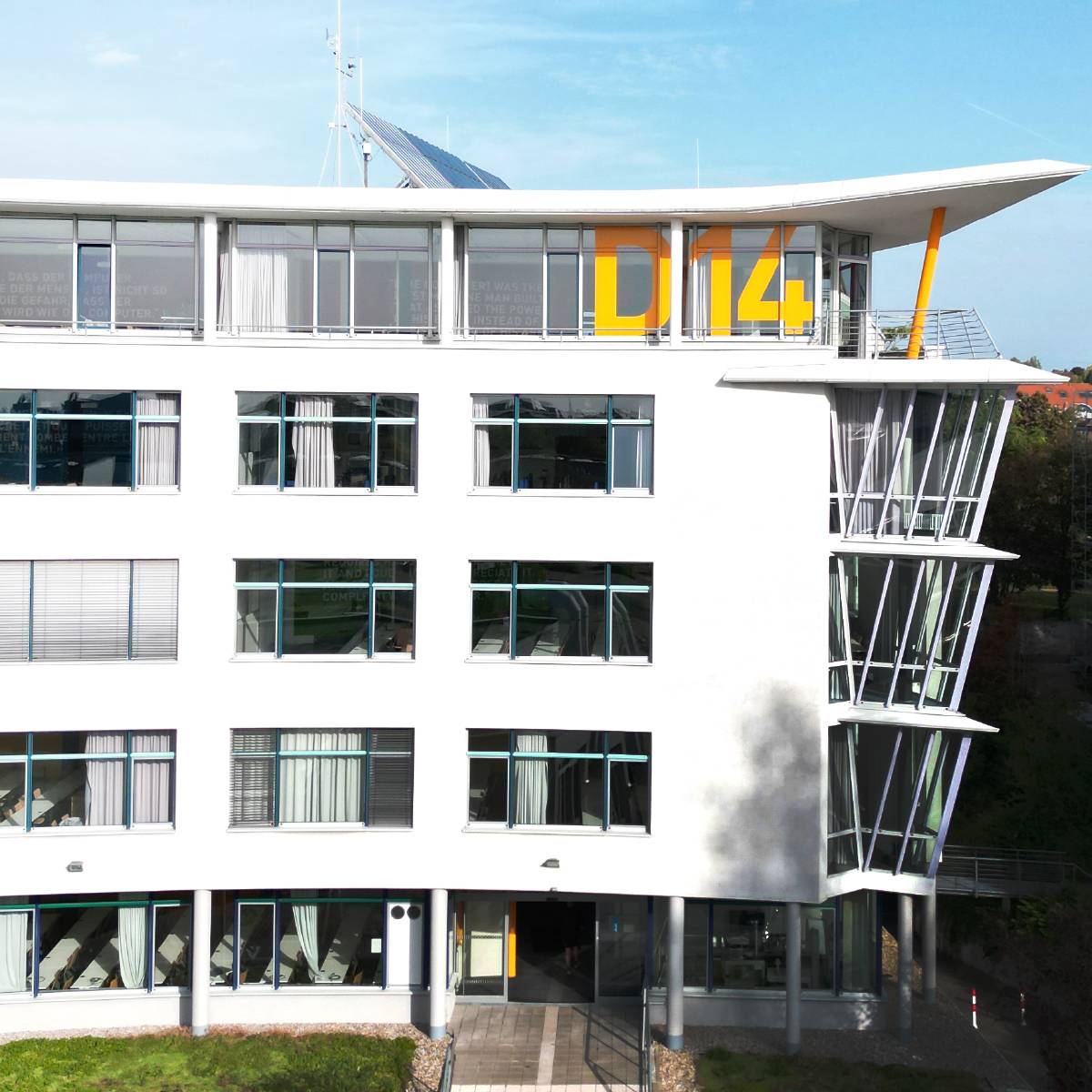 Modernes, weißes Bürogebäude mit vielen Fenstern und einer auffällig gelben Beschriftung „D14“ im obersten Stockwerk.