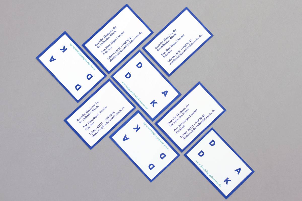 Acht weiße Visitenkarten mit blauem Rand liegen auf grauem Hintergrund in einer diagonalen Anordnung; auf einigen Karten sind Kontaktdaten und auf anderen blaue Buchstaben sowie Symbole zu sehen.