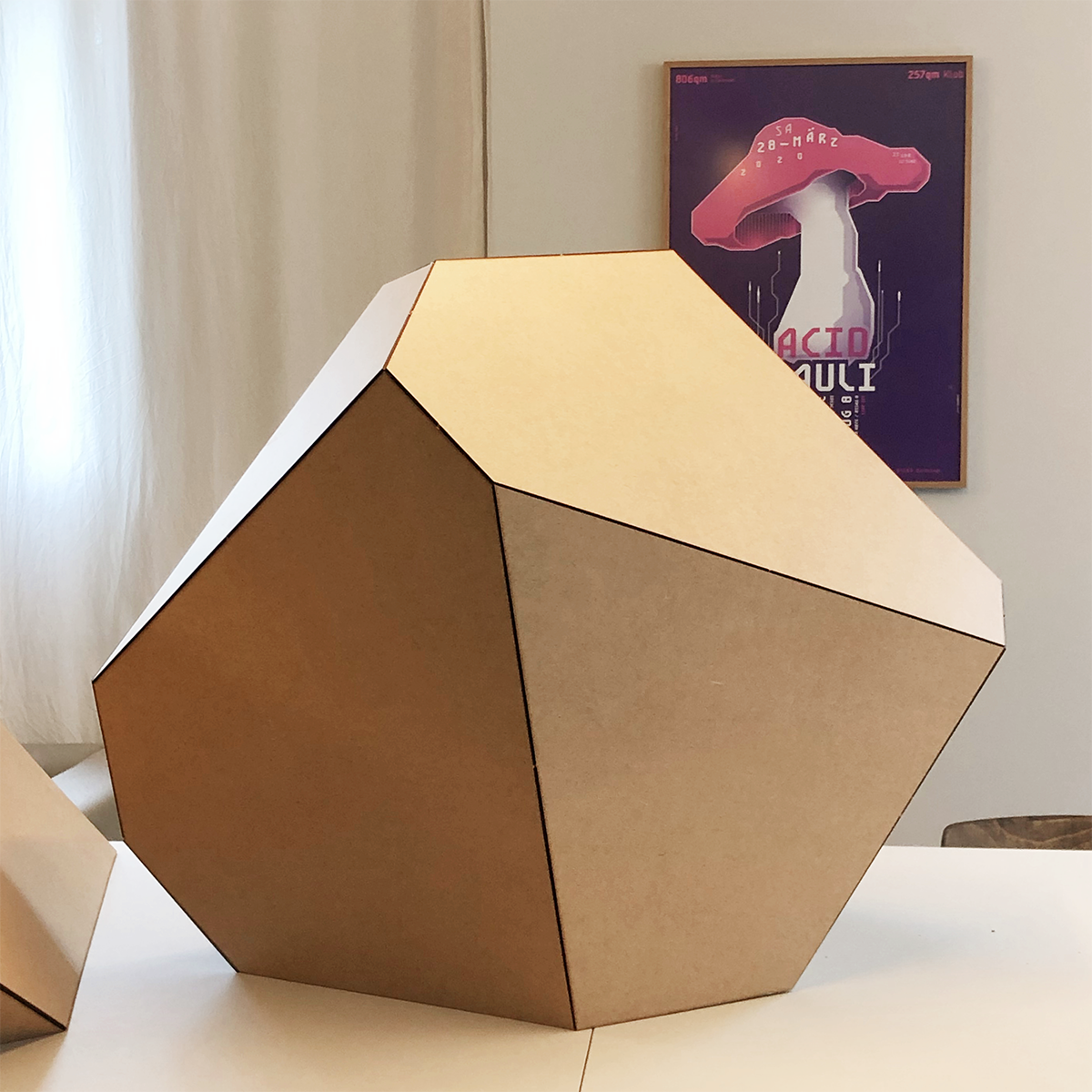 Ein großer, geometrischer Polyeder aus Holz steht auf einem Tisch; im Hintergrund hängt ein Poster mit einem Pilzmotiv.