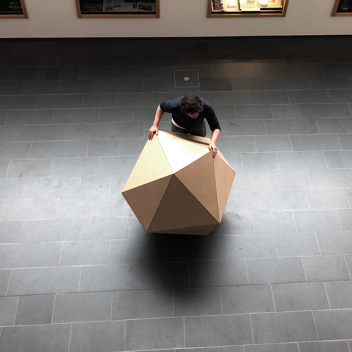 Person betrachtet ein großes, geometrisches, zwölfflächiges Objekt auf einem grauen Fliesenboden von oben.