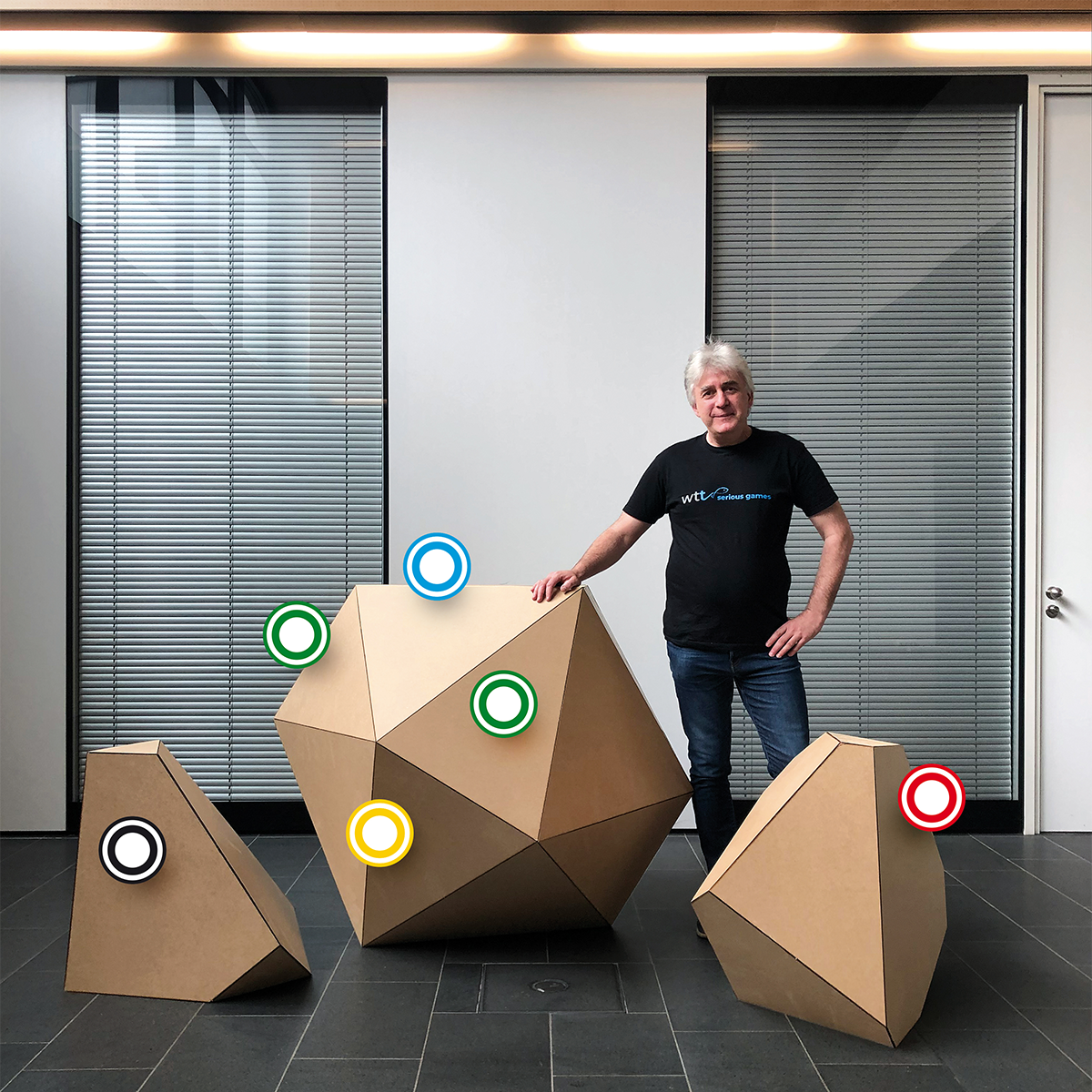 Ein Mann steht in einem modernen Innenraum neben drei großen, geometrischen Pappskulpturen mit bunten Kreismarkierungen.