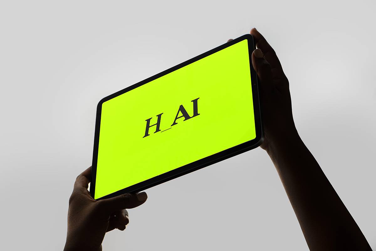 Zwei Hände halten ein Tablet mit leuchtend gelbem Bildschirm, auf dem in schwarzer Schrift „H_AI“ steht.